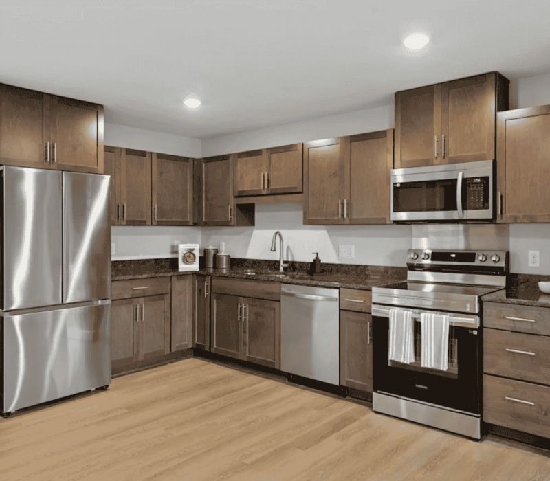Modern kitchen with brown cabinets, stainless steel appliances, and wood flooring under bright lighting.