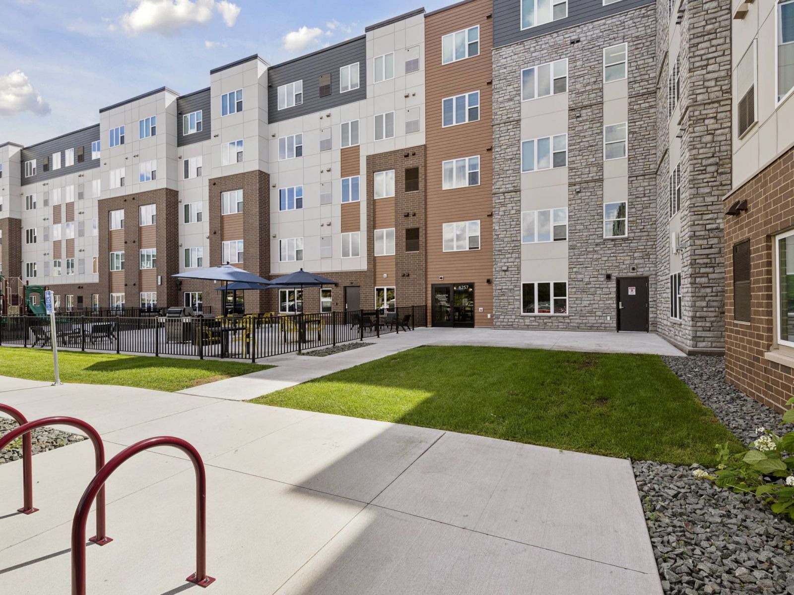 Modern apartment building with outdoor seating area, playground, and bike racks on a sunny day.