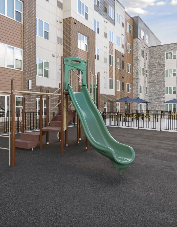 Green slide and climbing playground on blacktop, surrounded by fencing, near modern apartment buildings.
