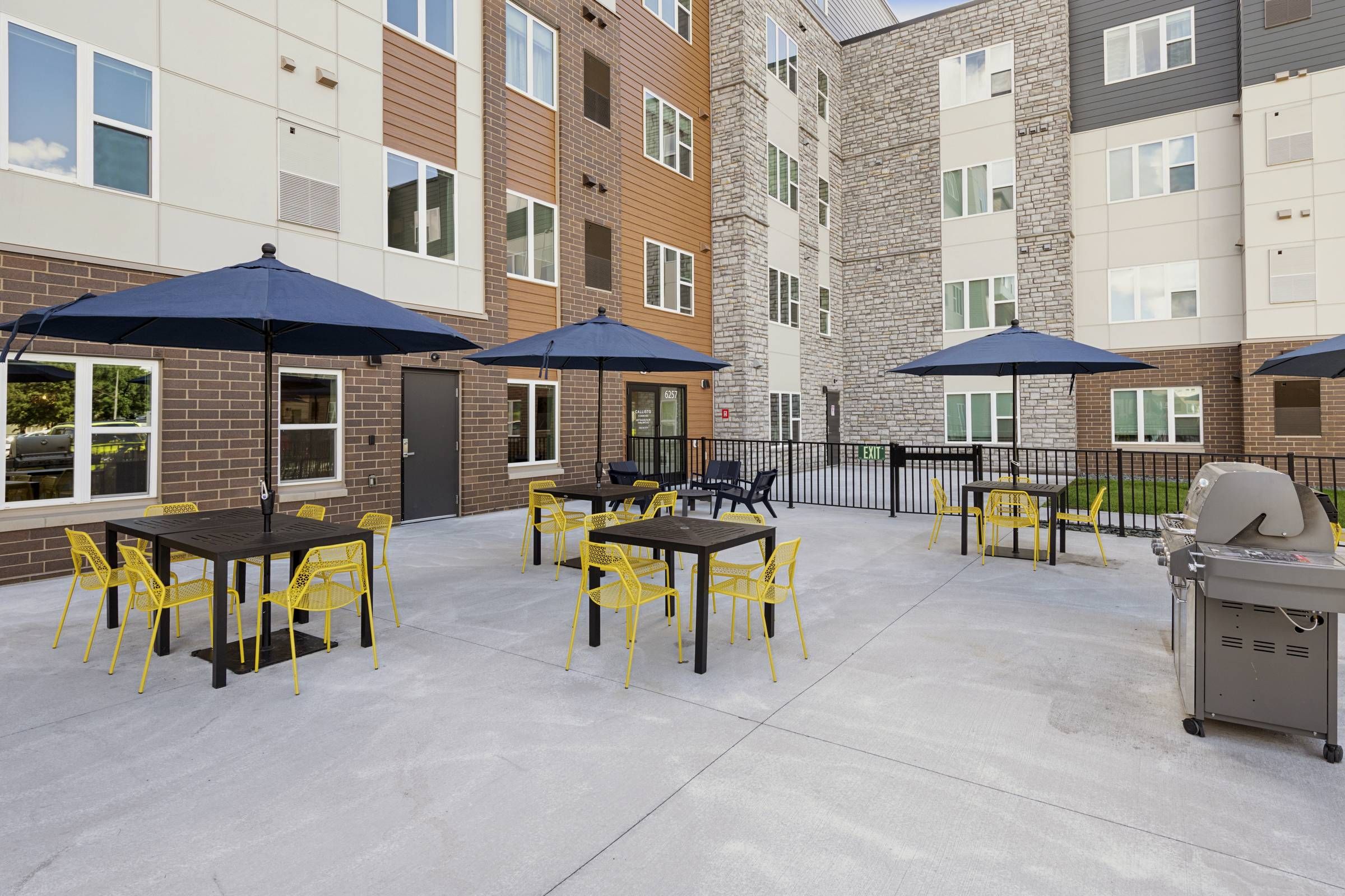 Outdoor patio with tables, yellow chairs, black umbrellas, and a grill, surrounded by apartment buildings.