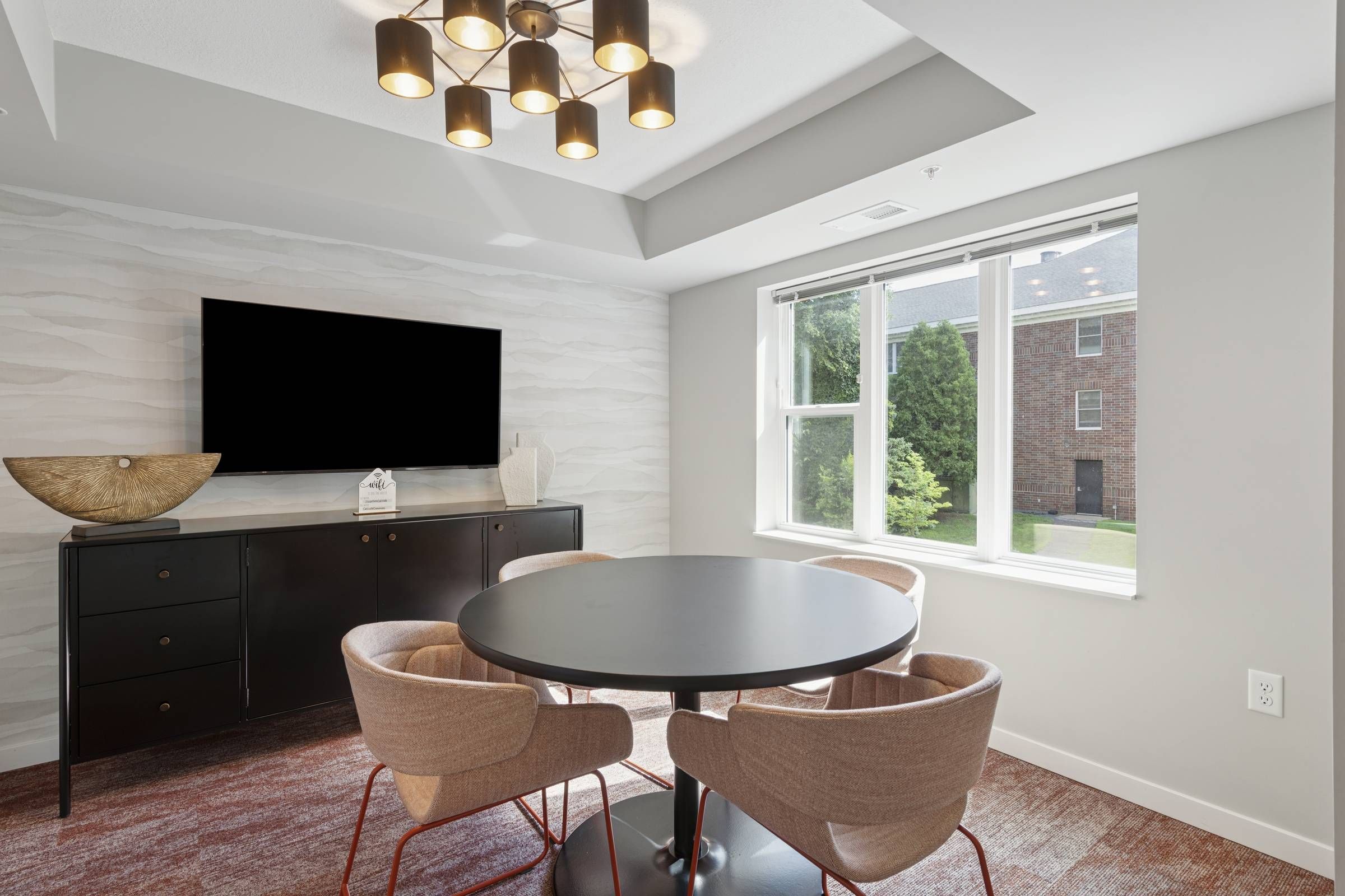 Modern meeting room with a round table, four chairs, TV, sideboard, and large window letting in natural light.