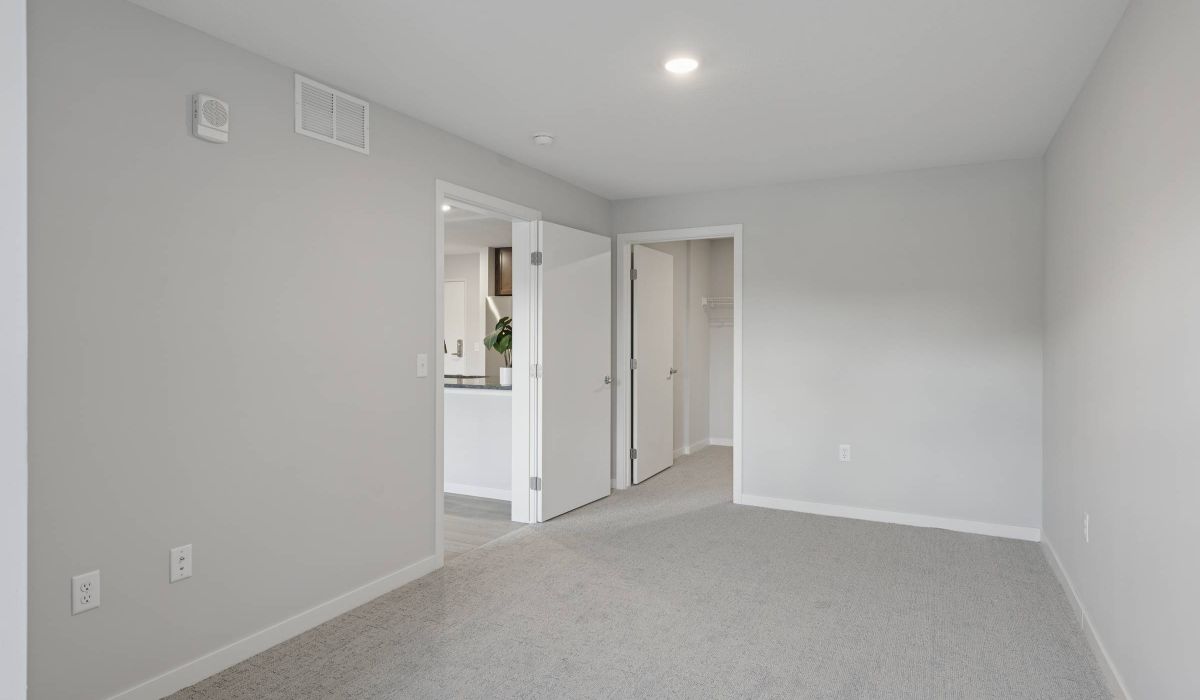 Empty, carpeted room with light gray walls, open doors, and view into a modern kitchen and closet.