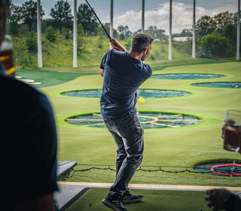 Man swings golf club at driving range with circular targets, people holding drinks in the foreground.