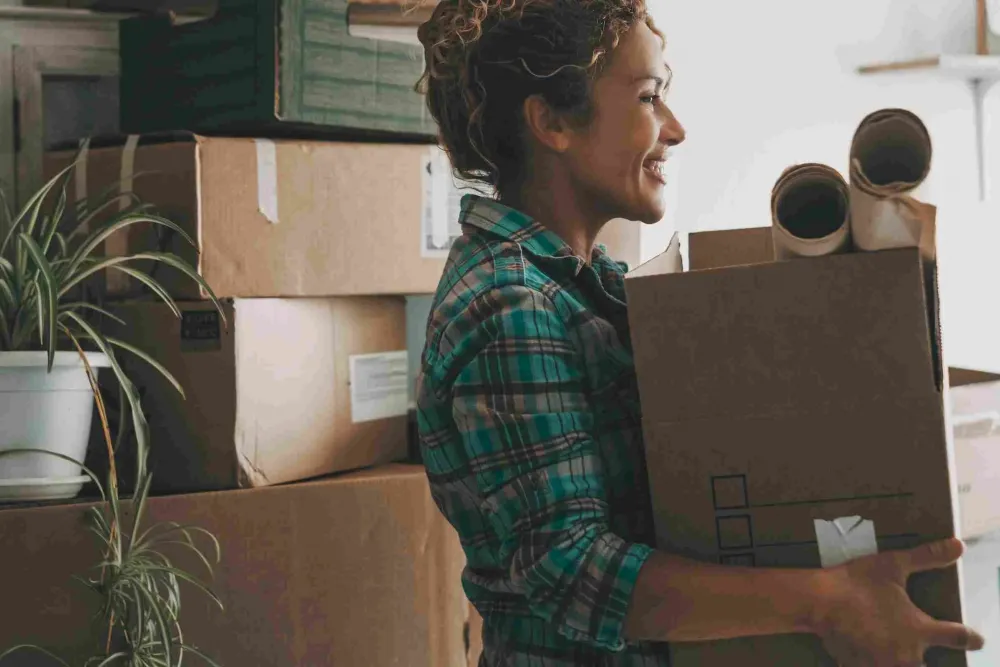 Smiling woman carrying a box and rolled papers, surrounded by moving boxes and a potted plant.