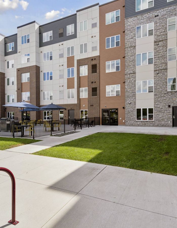 Modern apartment building with outdoor seating area, playground, and bike racks on a sunny day.