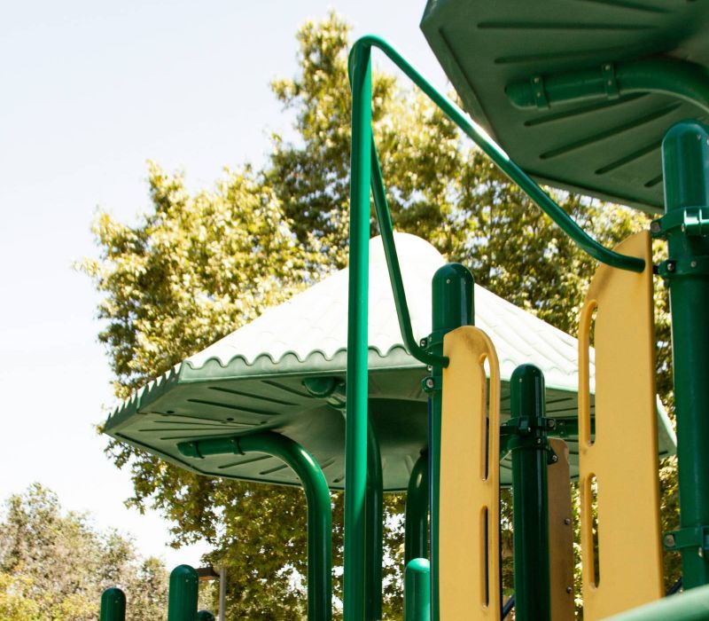A green and yellow playground structure with roofs, set outdoors near trees on a sunny day.
