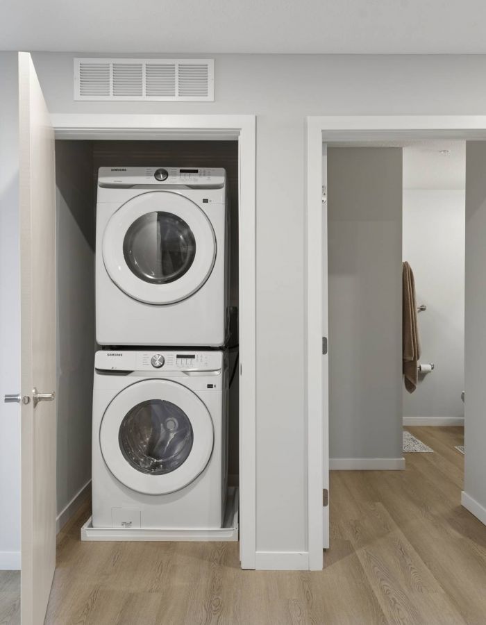 Stacked washer and dryer in a closet between two open doors in a modern, light-filled home hallway.