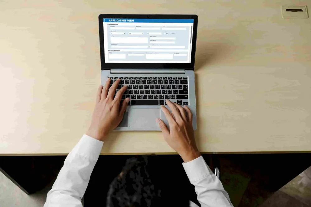 Person filling out an online application form on a laptop at a tidy desk, viewed from above.