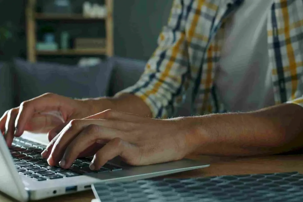 Person typing on a laptop at a desk, wearing a plaid shirt, with a blurred background.