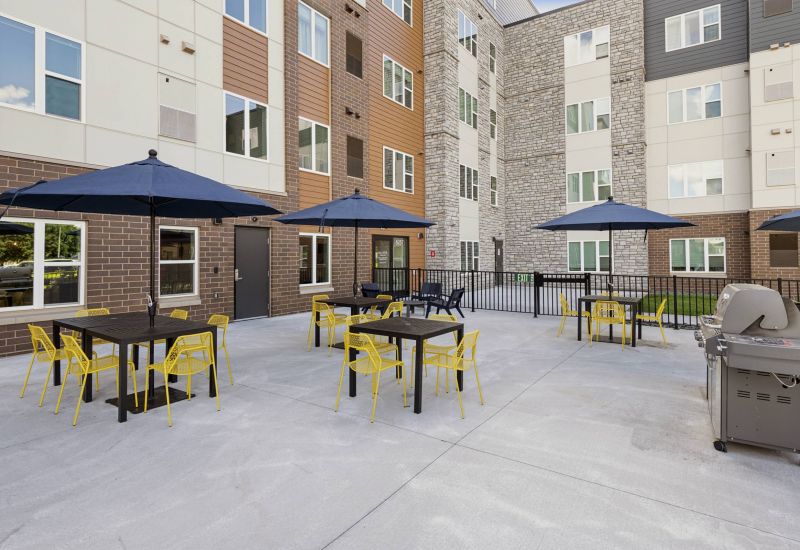 Outdoor patio with tables, yellow chairs, black umbrellas, and a grill, surrounded by apartment buildings.