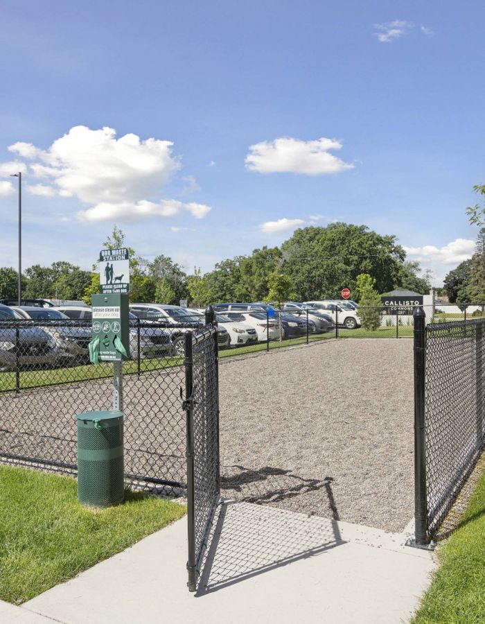 Fenced dog park with gravel ground, waste station, and parked cars in the background on a sunny day.